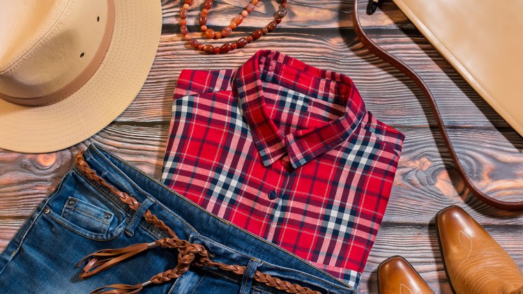 Rotes kariertes Hemd, blaue Jeans mit braunem Gürtel, beiger Hut, braune Stiefel, Perlenkette und Handtasche auf braunem Holzhintergrund.