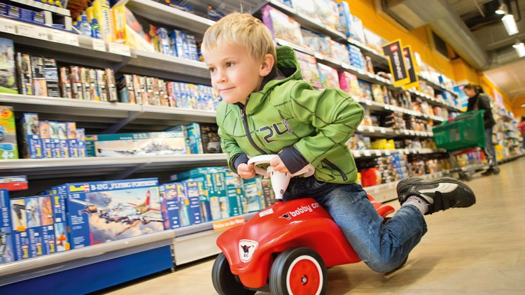 Kind in grüner Jacke fährt auf einem roten Bobby-Car durch eine Spielzeugabteilung mit Regalen voller Modellbaukästen.