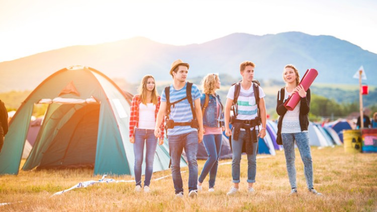 Fünf Personen mit Rucksäcken stehen auf einer Wiese vor Zelten in einer sonnigen Landschaft mit Bergen im Hintergrund.