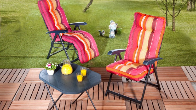 Zwei Gartenstühle mit rot-gelb gestreiften Polstern und ein Tisch mit Orangensaft und Blumen stehen auf einer Terrasse vor einer Wiese.
