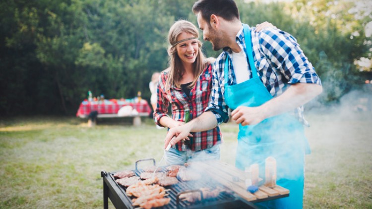 Person in blauem Grill-Kochhemd grillt Fleisch und Gemüse auf einem Holzkohlegrill im Freien, im Hintergrund Wiese und Bäume.