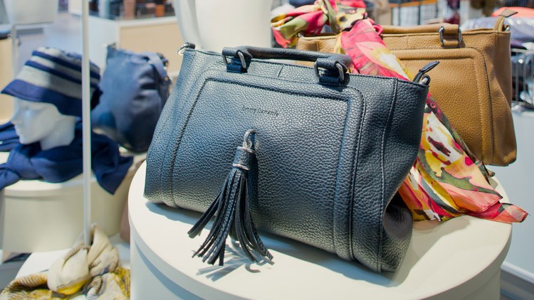 Schwarze Handtasche mit Fransenanhänger auf weißem Tisch, daneben Tücher, eine braune Tasche und ein blaues Accessoire.