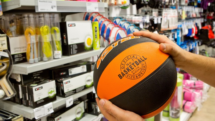 Person hält einen orange-schwarzen Basketball vor einem Regal mit Sportartikeln wie Springseilen und Tischtennisschlägern.