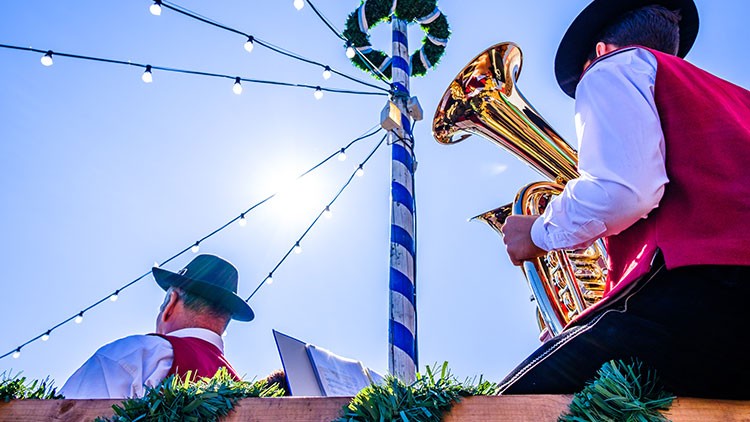 Zwei Musiker in traditionellen Westen spielen vor einem geschmückten Maibaum Tuba und Trompete unter freiem Himmel.