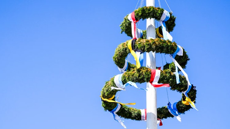 Ein weißer Maibaum mit mehreren grünen Kränzen und bunten Stoffbändern steht vor einem klaren blauen Himmel.