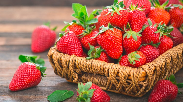 Ein geflochtener Korb voller roter Erdbeeren mit grünen Blättern steht auf einem Holztisch, einzelne Beeren liegen daneben.