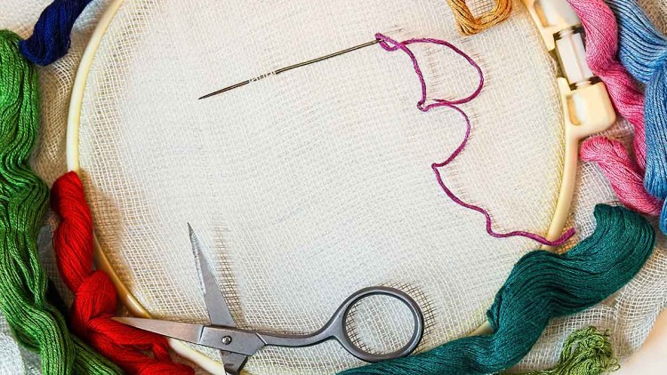 Stickrahmen mit feinem Stoff, eingebundene Nadel mit violettem Faden, bunte Garnfäden und kleine Schere liegen daneben.