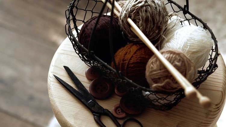 Drahtkorb mit Wollknäueln und Stricknadeln steht auf einem hellen Holzhocker, daneben liegen Knöpfe und eine schwarze Schere.