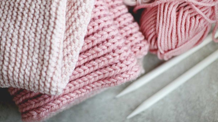 Weiche, rosafarbene Strickstücke liegen neben Knäueln rosafarbener Wolle und zwei weißen Stricknadeln auf grauem Untergrund.
