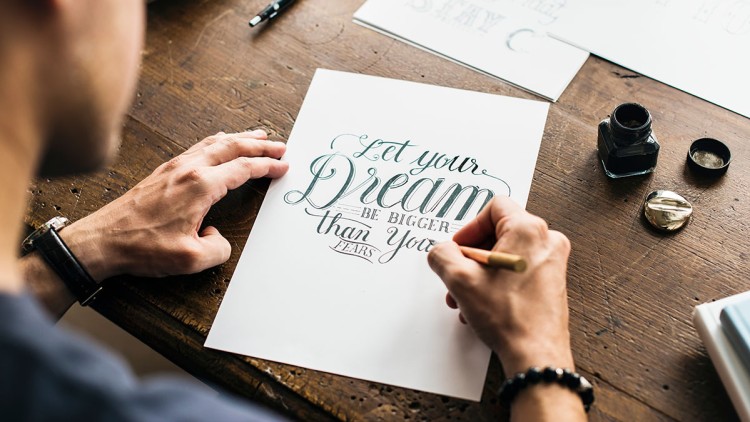 Eine Person schreibt mit Bleistift auf Papier den Spruch „Let your dream be bigger than your fears“, daneben Tinte und ein Glas.