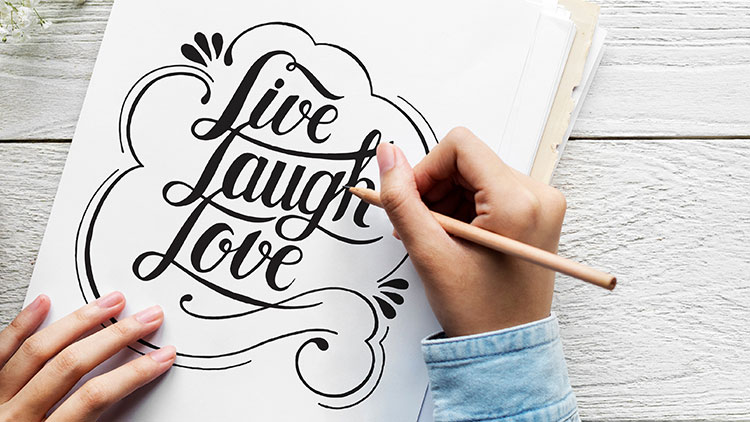 Eine Hand hält einen Bleistift und schreibt die Worte Live, Laugh, Love in geschwungener Schrift auf weißes Papier.