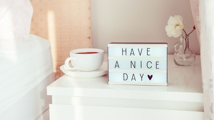 Eine weiße Nachttischlampe mit der Aufschrift HAVE A NICE DAY und einer Tasse Kaffee daneben auf einem weißen Nachtschrank.