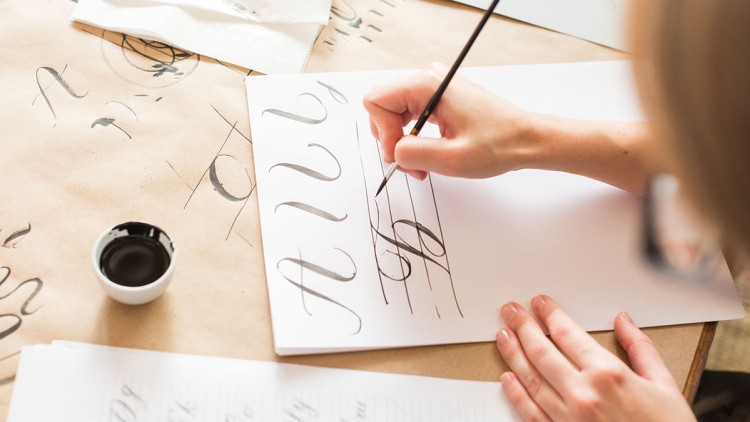 Eine Hand malt mit einem Pinsel Buchstaben in schwarzer Tinte auf ein weißes Blatt mit vorgegebenen Linien.