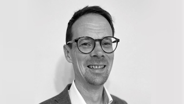 Eine Person mit kurzem Haar trägt eine Brille, ein helles Hemd und ein Sakko vor neutralem Hintergrund.