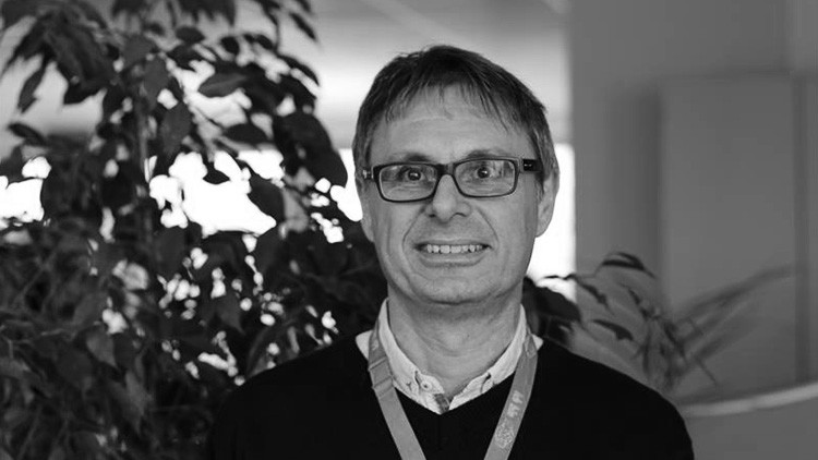 Person mit hellem Hemd und dunklem Pullover trägt ein Schlüsselband, im Hintergrund sind Büropflanzen erkennbar.