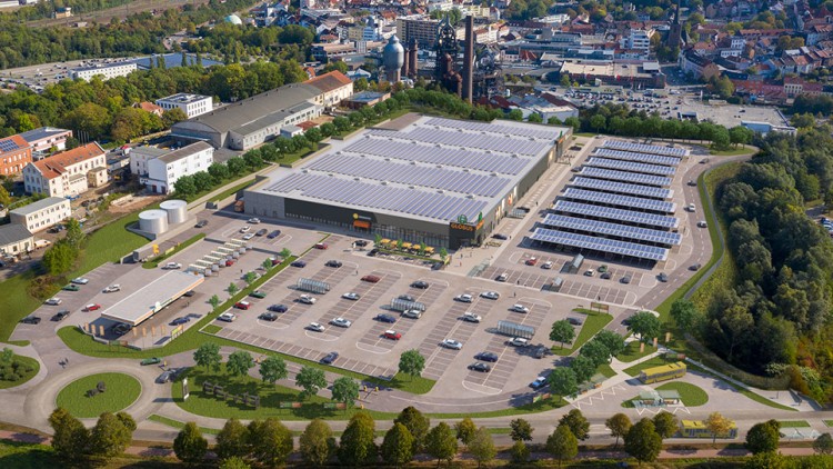 Luftaufnahme eines GLOBUS Marktes mit Photovoltaikanlagen auf dem Dach, großem Parkplatz und umliegender grüner Landschaft.