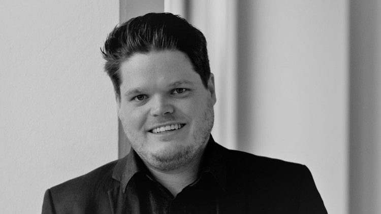 Person in schwarzem Sakko und schwarzem Hemd steht vor einer weißen Wand und einem Türrahmen.