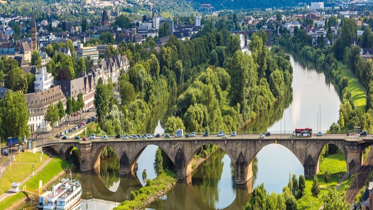Eine steinerne Brücke mit Bögen und Verkehr führt über einen Fluss, umgeben von Bäumen und einer Stadt im Hintergrund.