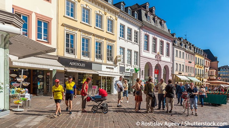 Mehrere Personen gehen oder stehen auf einem Platz vor pastellfarbenen Gebäuden und Geschäften bei sonnigem Wetter.
