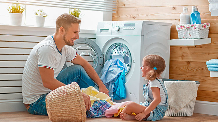 Ein Kind sitzt mit Spielzeug vor einer geöffneten Waschmaschine, daneben ein Korb mit Wäsche, Holzverkleidung im Hintergrund.