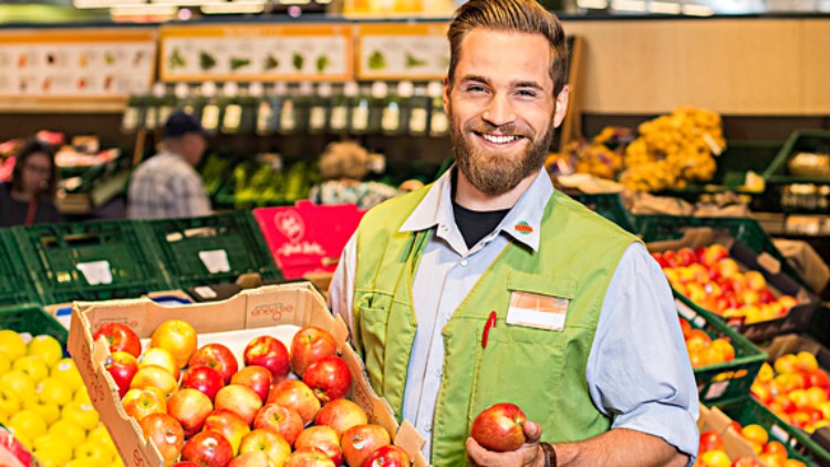 Eine Person in grüner Weste hält eine Apfelkiste, im Hintergrund Obstkisten und eine Preistafel mit Apfelsorten.