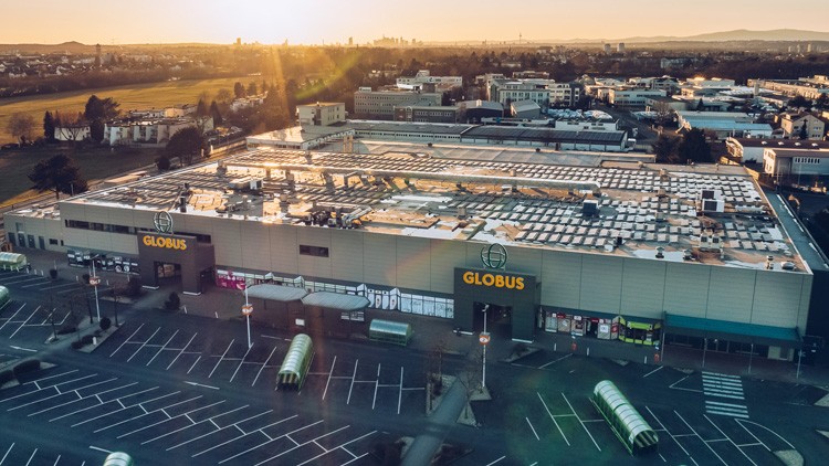 Das GLOBUS Gebäude mit großem Parkplatz und leeren Parkflächen, aufgenommen aus der Luft bei Sonnenuntergang.