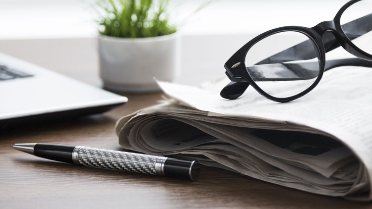 Gefaltete Zeitung, schwarze Brille und ein silber-schwarzer Kugelschreiber liegen auf einem Tisch neben einem Laptop und einer Pflanze.