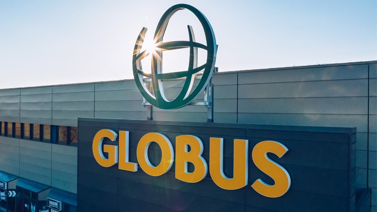 Das GLOBUS-Logo mit grünem Globus-Symbol und gelbem Schriftzug thront auf einem Gebäude vor blauem Himmel mit Sonnenstrahlen.