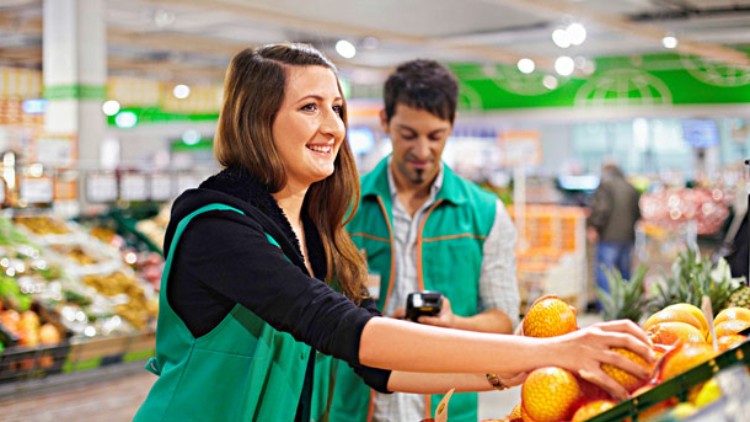 Person in grüner Arbeitsweste legt Orangen in ein Regal, im Hintergrund ein weiteres Regal mit Gemüse und Obst.