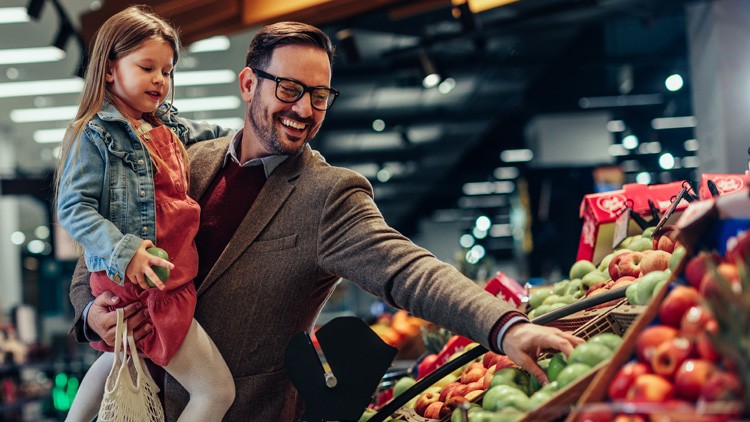 Eine Person trägt ein Kind und greift nach grünem Obst in einem Supermarkt mit beleuchteten Obst- und Gemüseständen.