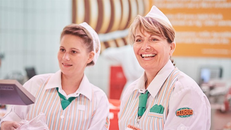 Zwei Personen in gestreiften GLOBUS-Uniformen mit grünen Krawatten stehen an einer Kasse in einem Markt.