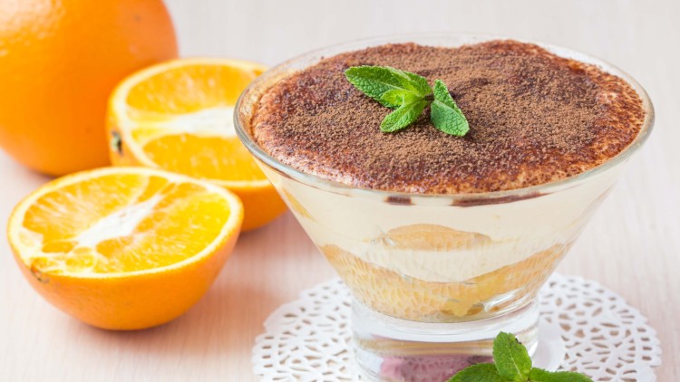 Glasdessert mit Kakaopulver und Minzblättern dekoriert, daneben halbe und ganze Orangen auf einem hellen Tisch.