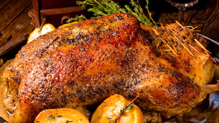 Goldbraune gebratene Gans mit Kräutern und gebackenen Apfelhälften in einem schwarzen Bräter auf Holztisch.