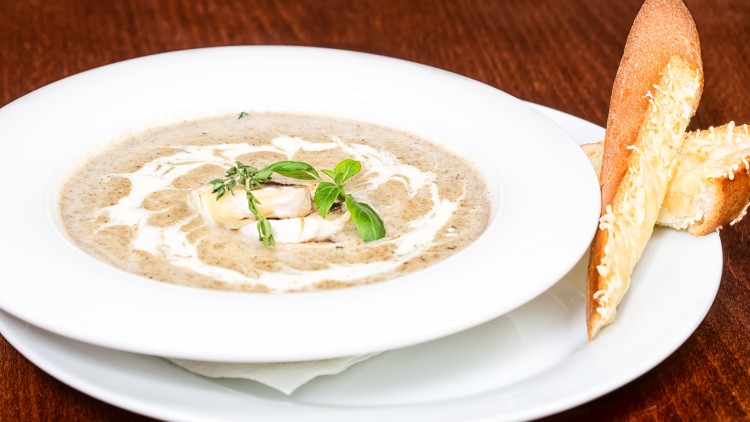 Cremige braune Suppe mit Sahne, Kräutergarnitur und Brot mit geschmolzenem Käse auf einem weißen Teller.