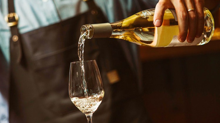 Eine Person in Schürze schenkt Weißwein aus einer Flasche in ein transparentes Weinglas ein.