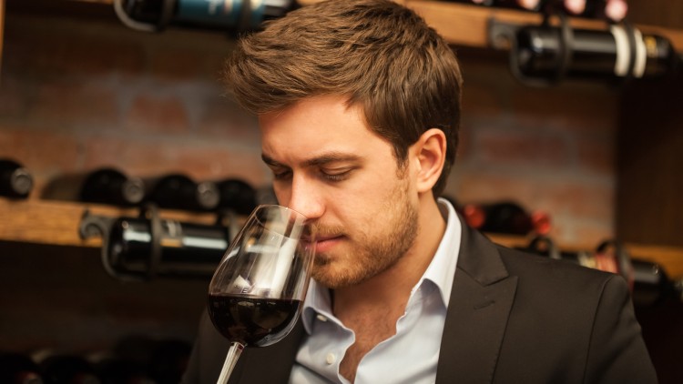 Eine Person in Anzug hält ein Glas Rotwein in der Hand, im Hintergrund ein Weinregal vor einer Ziegelwand.