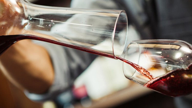 Rotwein wird aus einer durchsichtigen Karaffe in ein Weinglas gegossen, im Hintergrund verschwommene Person und Umgebung.