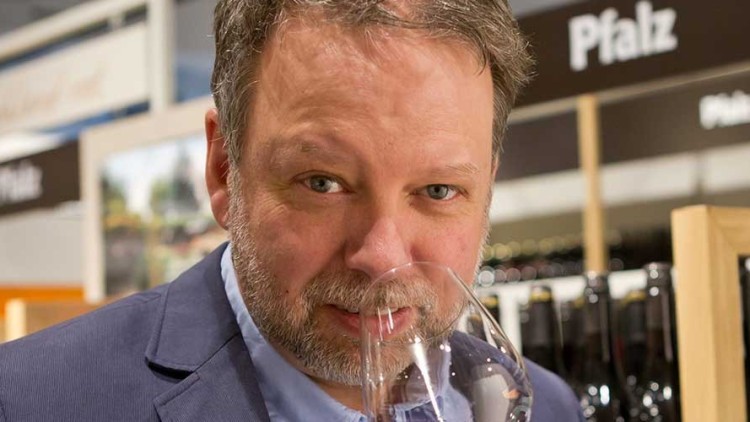 Weinregale mit Flaschen und dem Schriftzug Pfalz im Hintergrund, eine Person hält ein Weinglas in der Hand.
