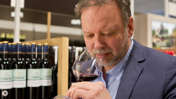 Regale mit Weinflaschen der Marke Weinbiet, eine Person hält ein Weinglas mit Rotwein in der Hand.