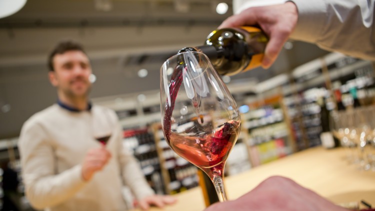 Eine Hand schenkt roten Wein aus einer Flasche in ein Weinglas ein, im Hintergrund stehen Regale mit Weinflaschen.