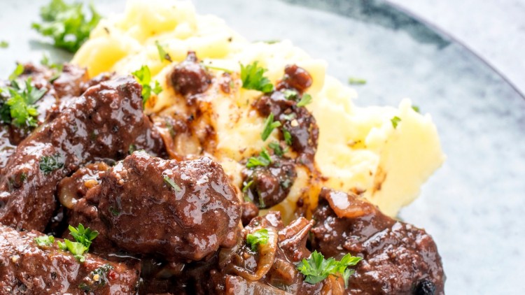 Dunkles Ragout mit Sauce und Petersilie neben gelbem Kartoffelpüree auf einem hellen Teller angerichtet.