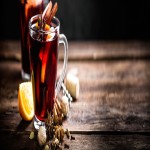 Zwei Gläser mit dunkelrotem Getränk, dekoriert mit Zimtstange und Orange, stehen auf einem Holztisch mit Gewürzen.