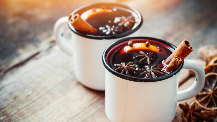 Zwei weiße Becher mit Glühwein, verziert mit Sternanis, Zimtstangen, Nelken und Orangenscheiben, auf einem Holztisch.