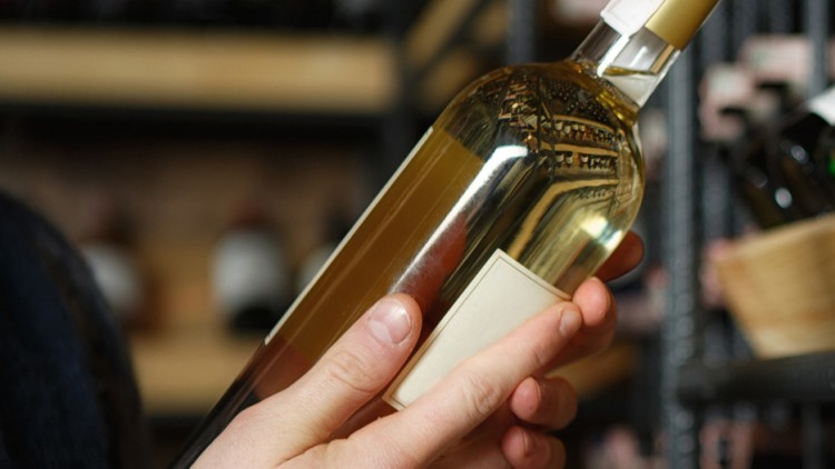 Eine Person hält eine Flasche Weißwein mit hellbeigem Etikett vor einem Regal mit weiteren Weinflaschen in der Hand.