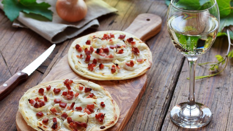 Zwei Flammkuchen mit Zwiebeln und Speck auf einem Holzbrett, daneben ein Glas Weißwein und ein großes Messer.