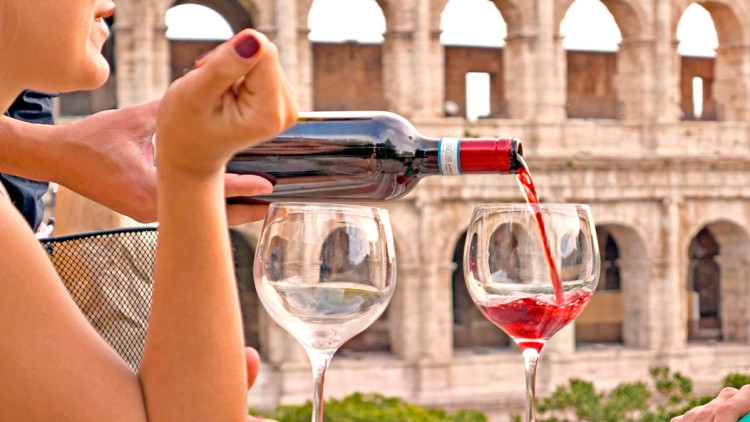 Eine Hand schenkt roten Wein aus einer Flasche in ein Weinglas ein, zwei Gläser und das Kolosseum sind im Hintergrund zu sehen.