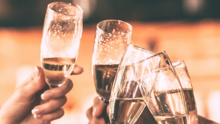 Mehrere Hände stoßen mit gläsernen Champagnerflöten an, im Hintergrund verschwommene Lichter in warmen Farbtönen.