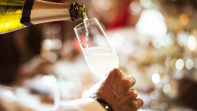 Ein helles Sektglas wird von einer Hand gehalten, während ein goldgelber Schaumwein aus einer Flasche hineingegossen wird.