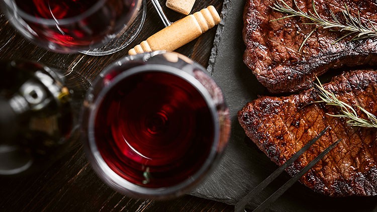 Zwei gegrillte Steaks mit Rosmarin und zwei Gläser Rotwein auf einem dunklen Untergrund mit Gebäckstangen.