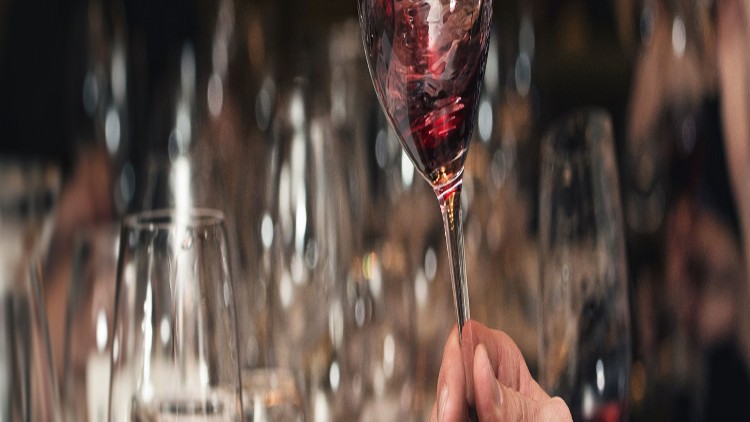 Eine Hand hält ein Weinglas mit rotem Wein, im Hintergrund unscharf mehrere Gläser und Personen.